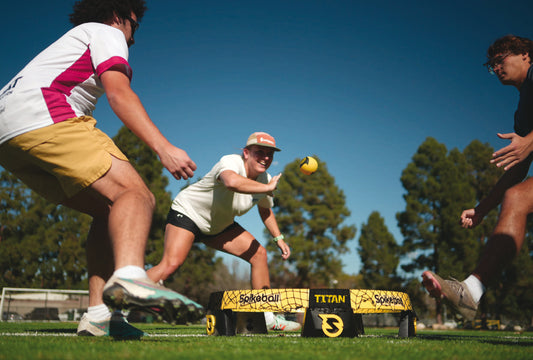 Two people playing Spikeball (titan set) with a ball on a grassy field, surrounded by trees and a clear blue sky.